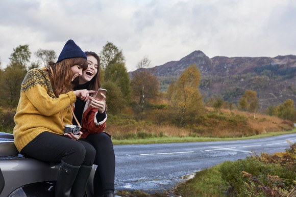 Two women laugh at something on their smartphones by a road out in the country on a cloudy day. 