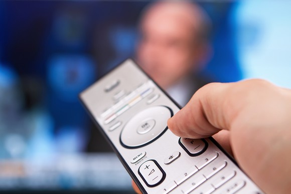 A hand holding a remote control with a TV in the background.