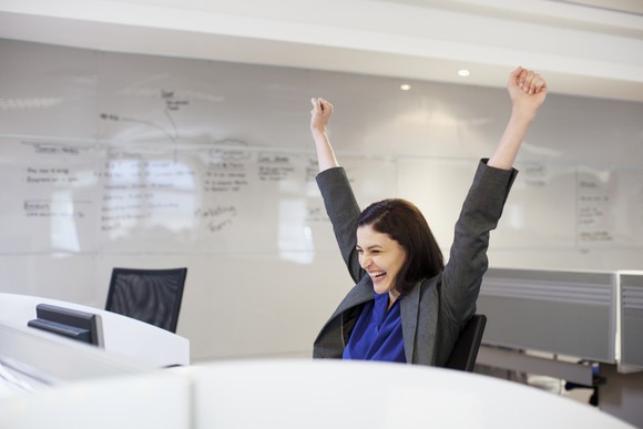 Woman sitting at a computer throwing her hands in the air and celebrating.