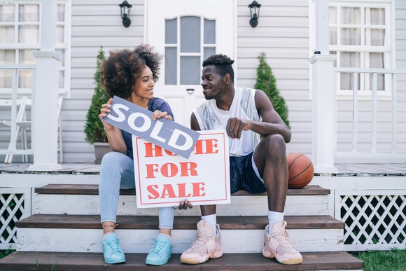 Two happy people sitting on the steps of their porch holding "For Sale" and "Sold" signs.