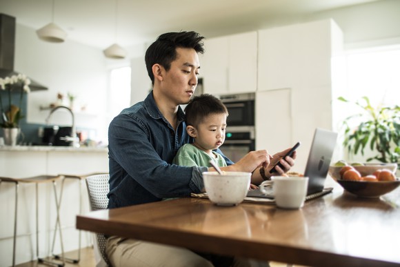 A father with his young son in his lap as he uses his smartphone in front of his laptop.