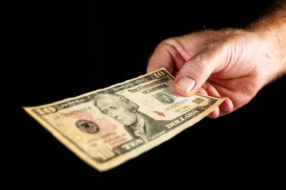 A man holding out a $10 bill with a dark background. 