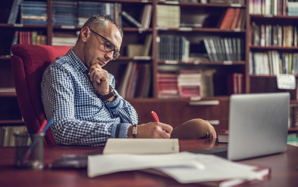 Man writing in notebook in study room with books and a laptop