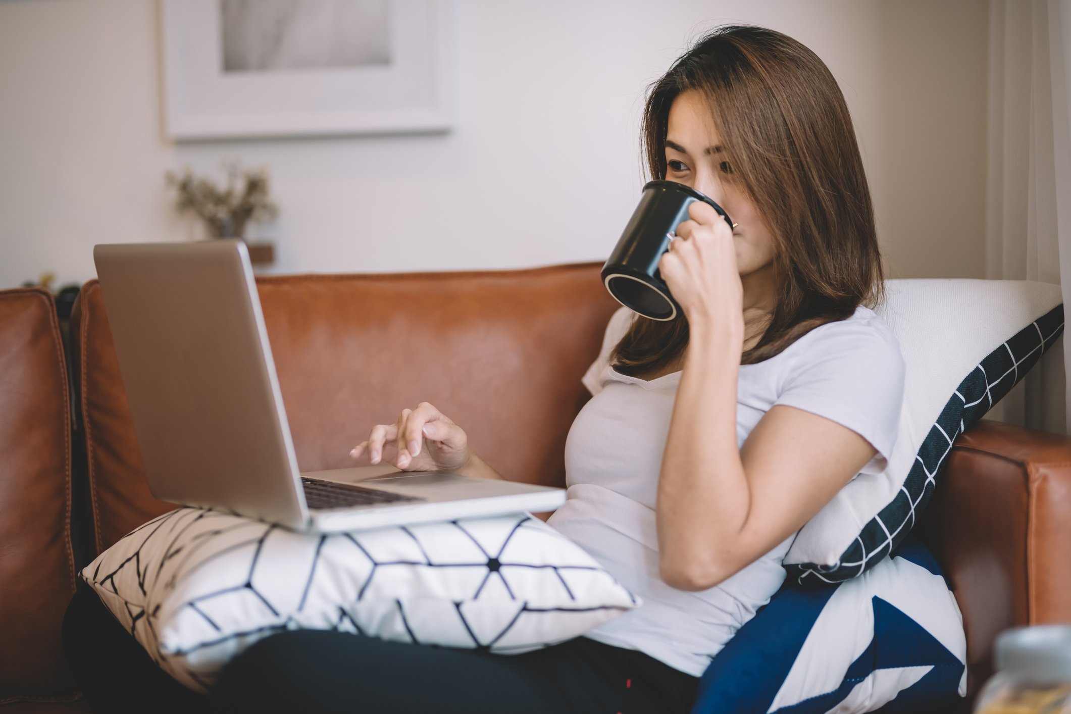 Woman sips coffee as she uses her laptop while lounging on her couch.
