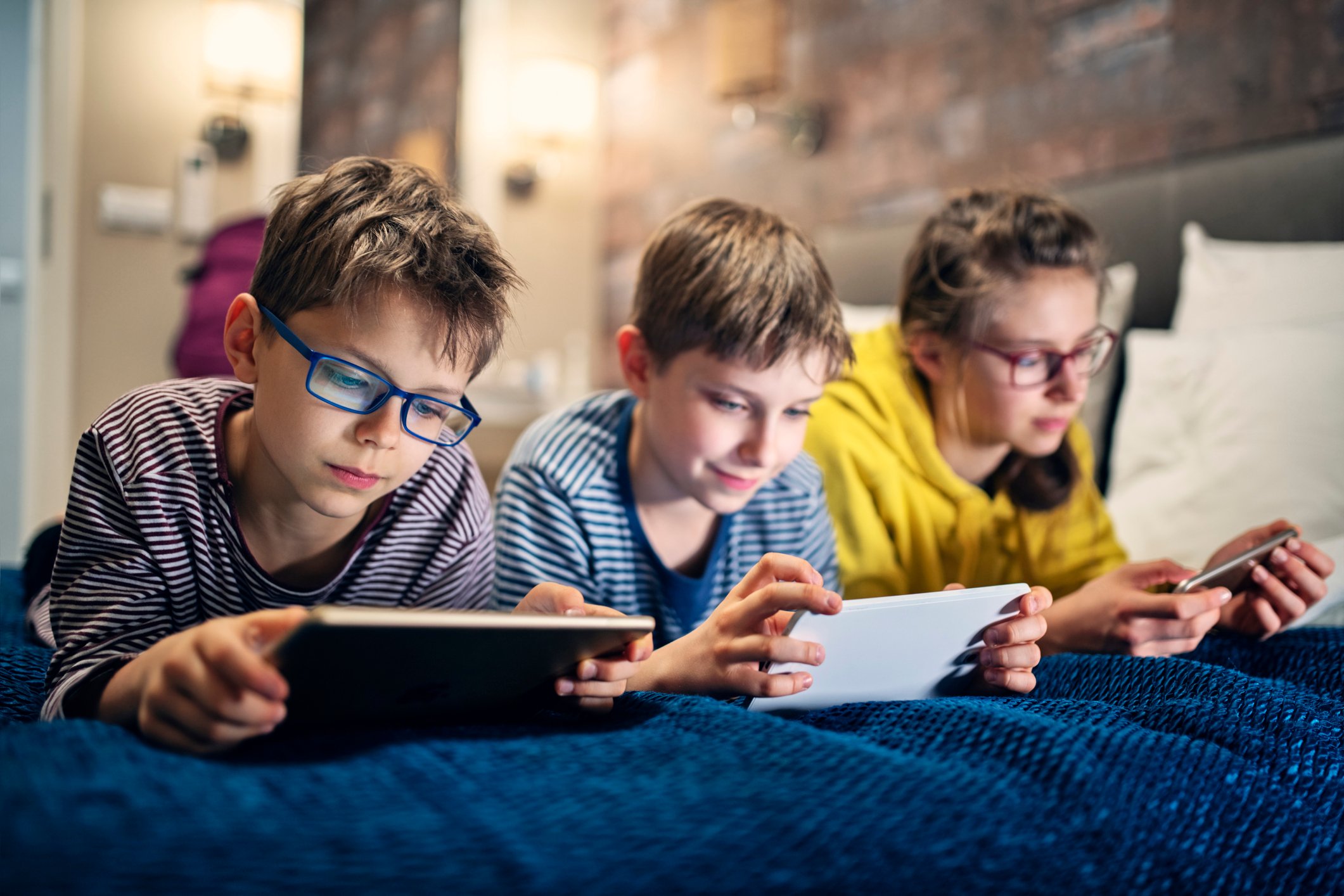 Three children play mobile games on a bed.