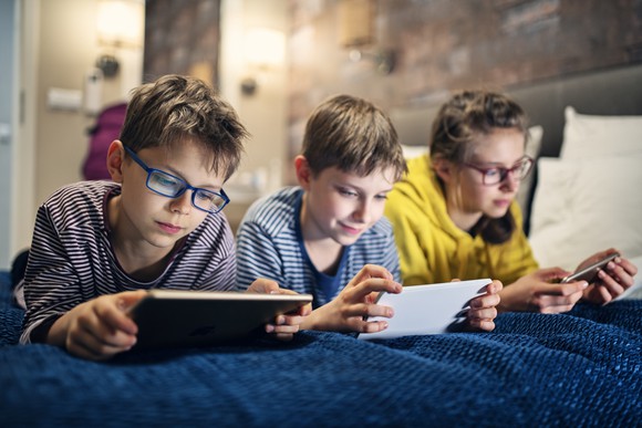 Three children play mobile games on a bed.