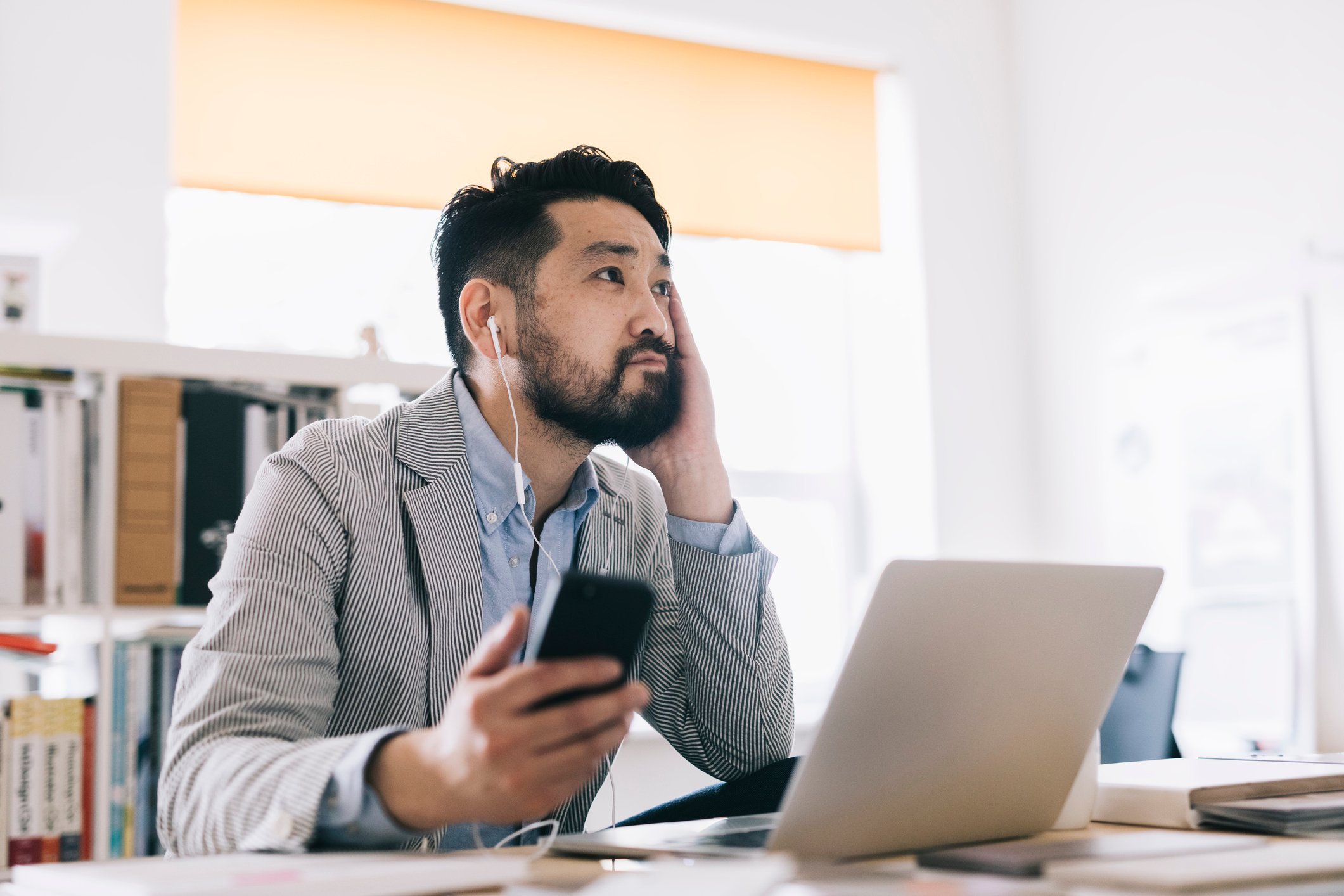 An Asian man listens to podcasts on his smartphone while sitting at a desk with a laptop atop it.