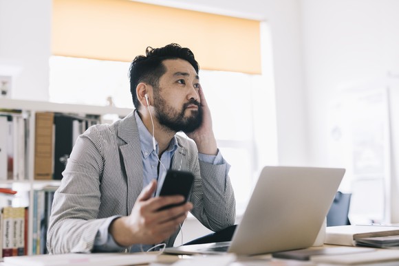 An Asian man listens to podcasts on his smartphone while sitting at a desk with a laptop atop it.