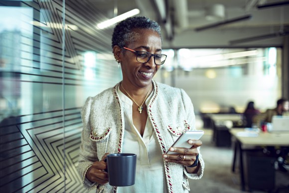 Senior woman looking at phone, holding a cup of coffee, smiling. 