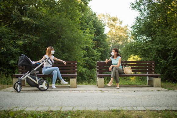 Two women sit six feet apart in masks on benches in a park, one with a stroller.