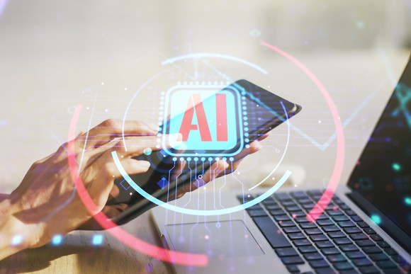 Overaly of letters AI over picture of hands on a smartphone before a laptop. 