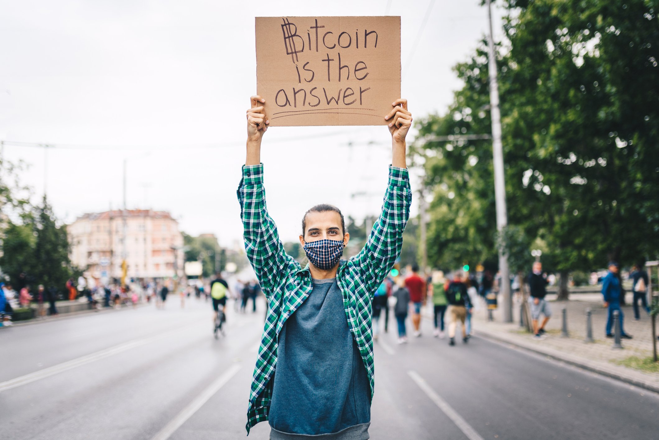 A masked man in the street holding a cardboard sign that reads "Botcoin is the answer."