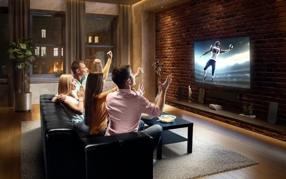 Two men and two women sitting on a couch and cheering as they watch a football game on a television