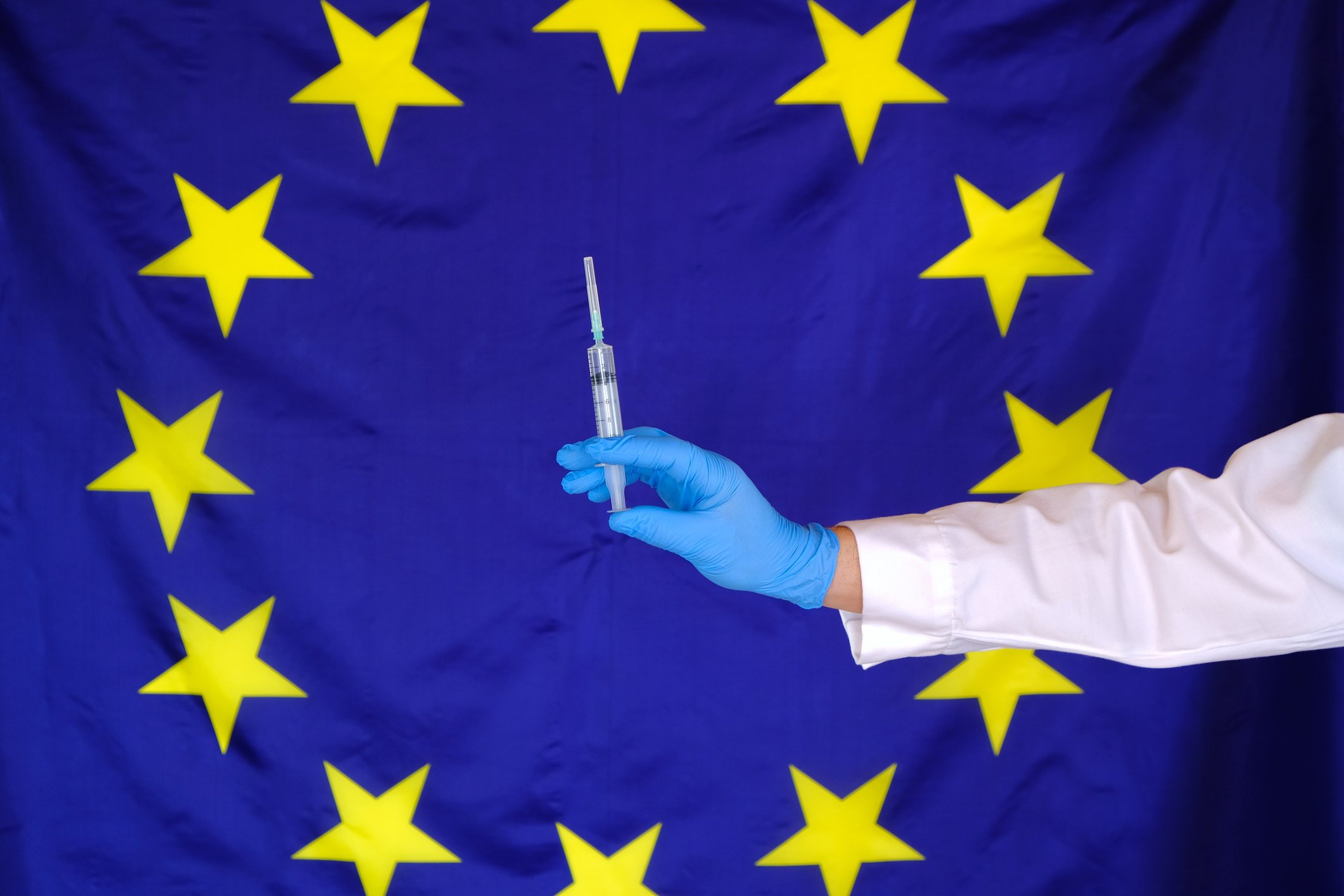 European Union flag with yellow stars in a circle on a blue background and a gloved hand holding a syringe with a needle in front of the flag