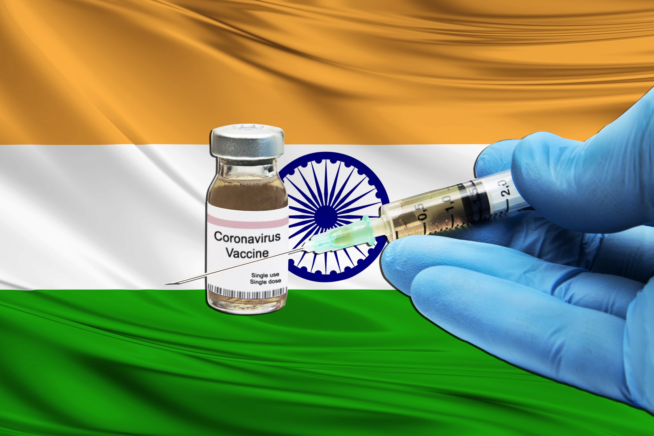 Gloved hand holding a syringe with a needle next to a coronavirus vaccine vial in front of an Indian flag