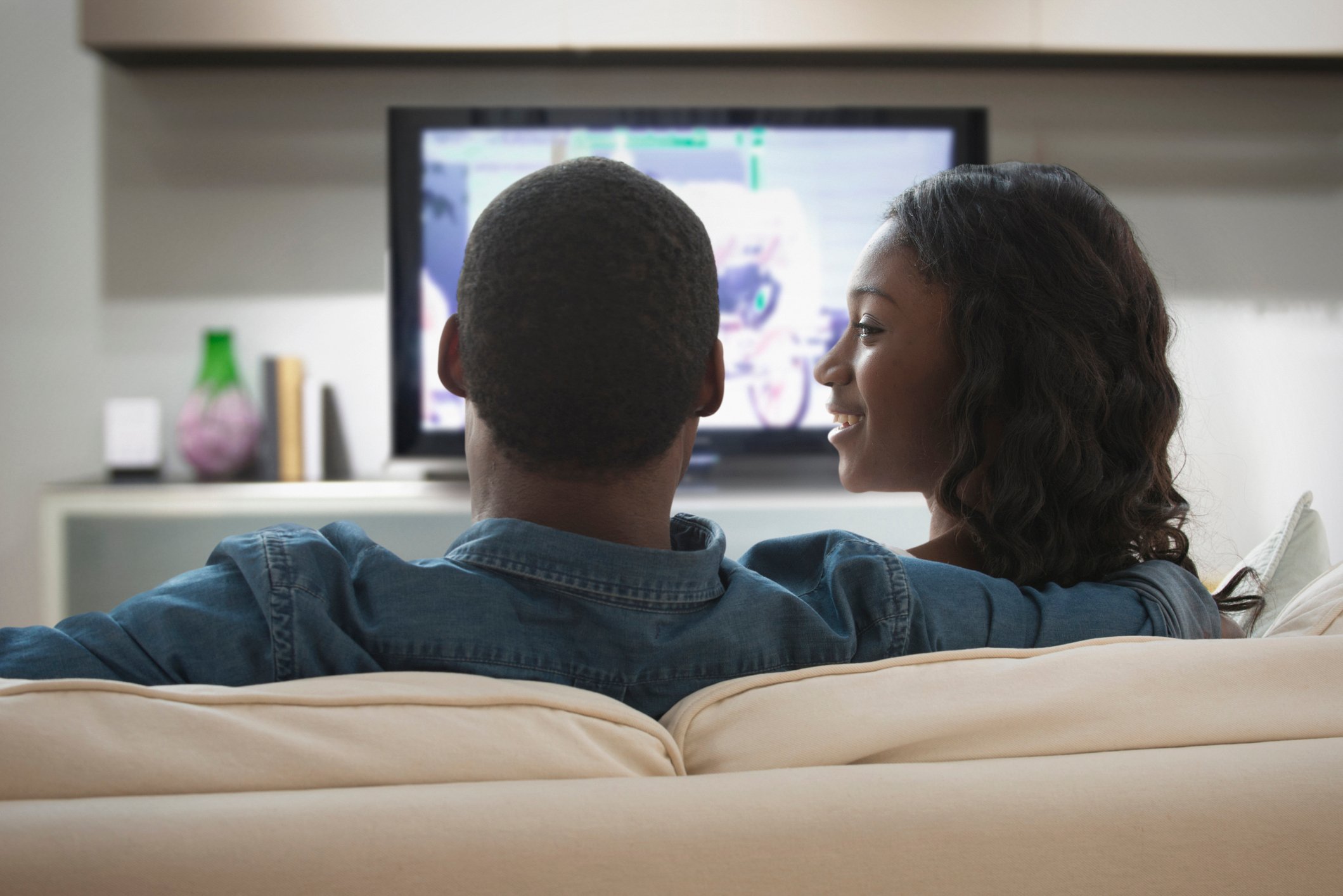A couple sits on a living room sofa watching television.