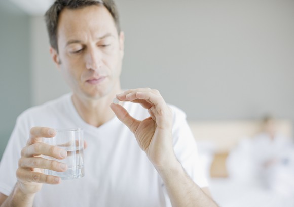 Person holds glass of water in one hand and a white pill in the other.