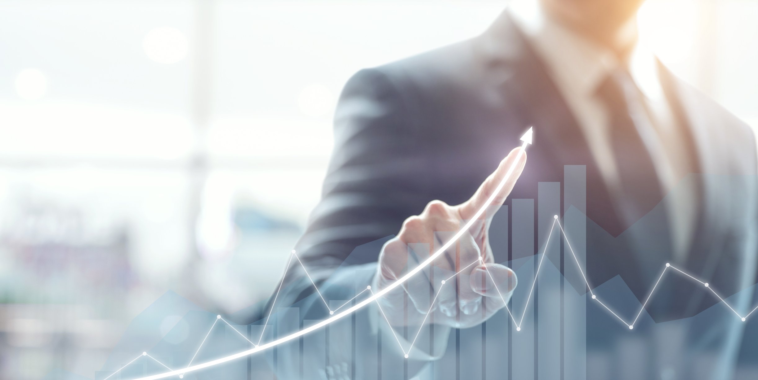 A businessman draws an upward arrow over a stock chart displayed on a transparent touchscreen.