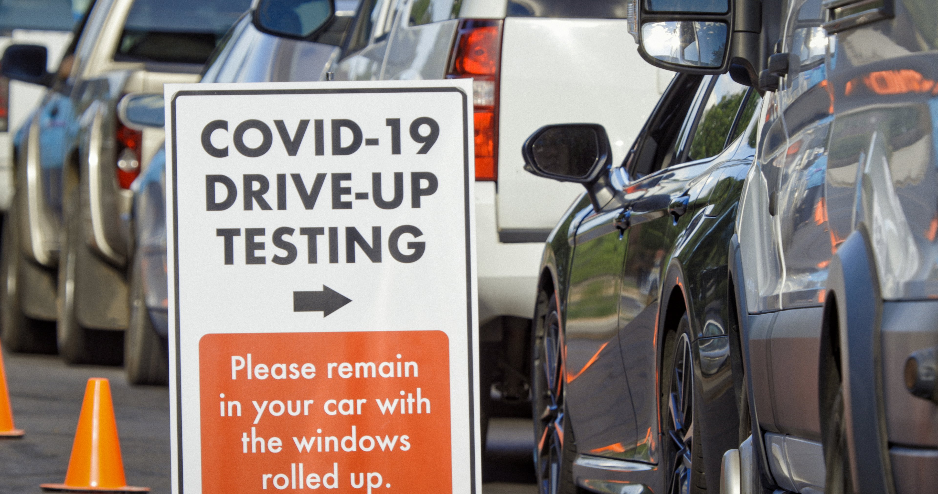 A COVID testing drive up location with cars lined up and an orange and white sign with an arrow pointing to the line.
