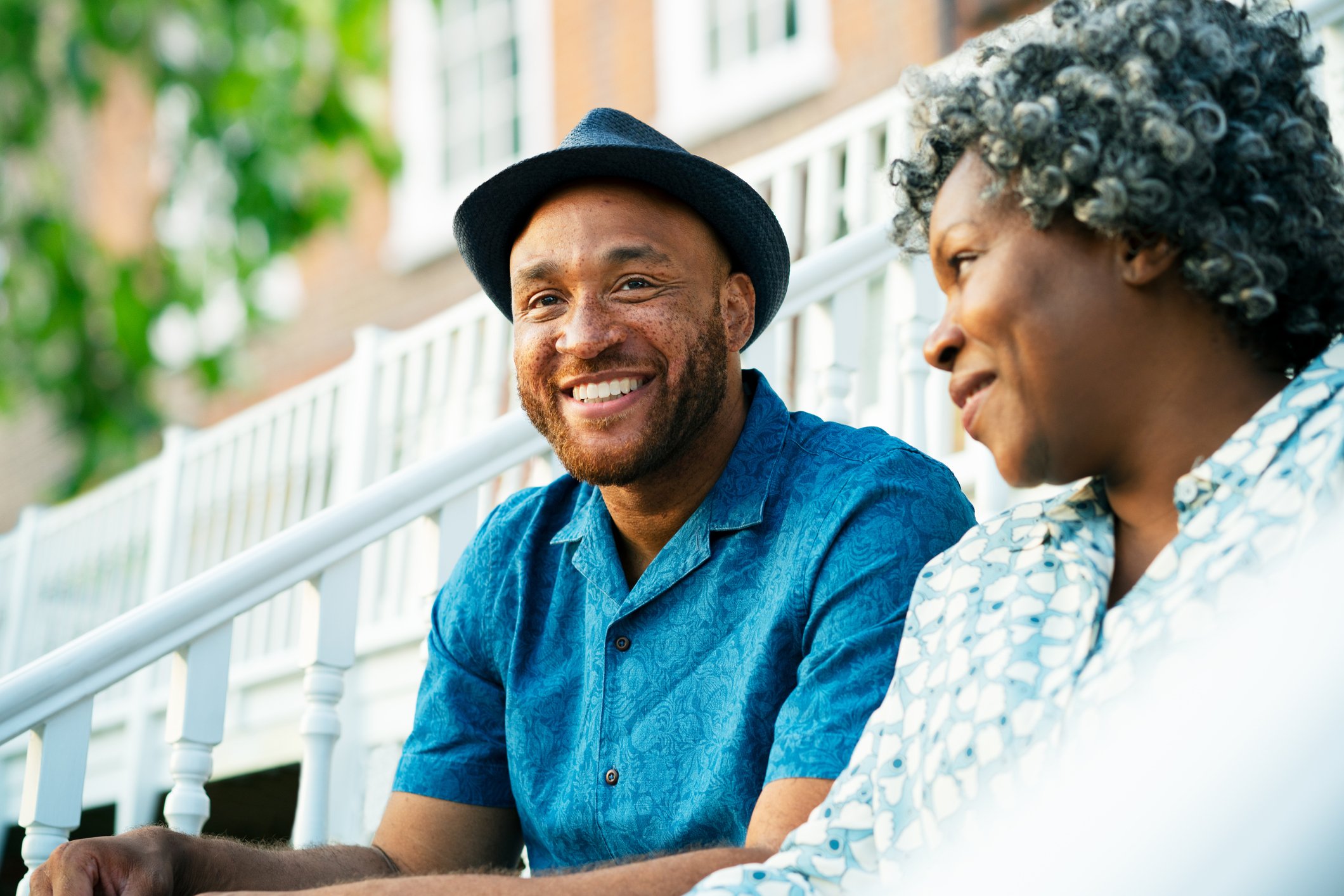 A man wearing a hat, smiling, sitting next to a woman, who is looking at him. 