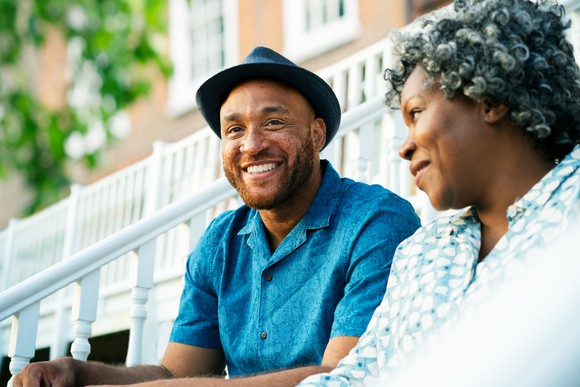 A man wearing a hat, smiling, sitting next to a woman, who is looking at him. 