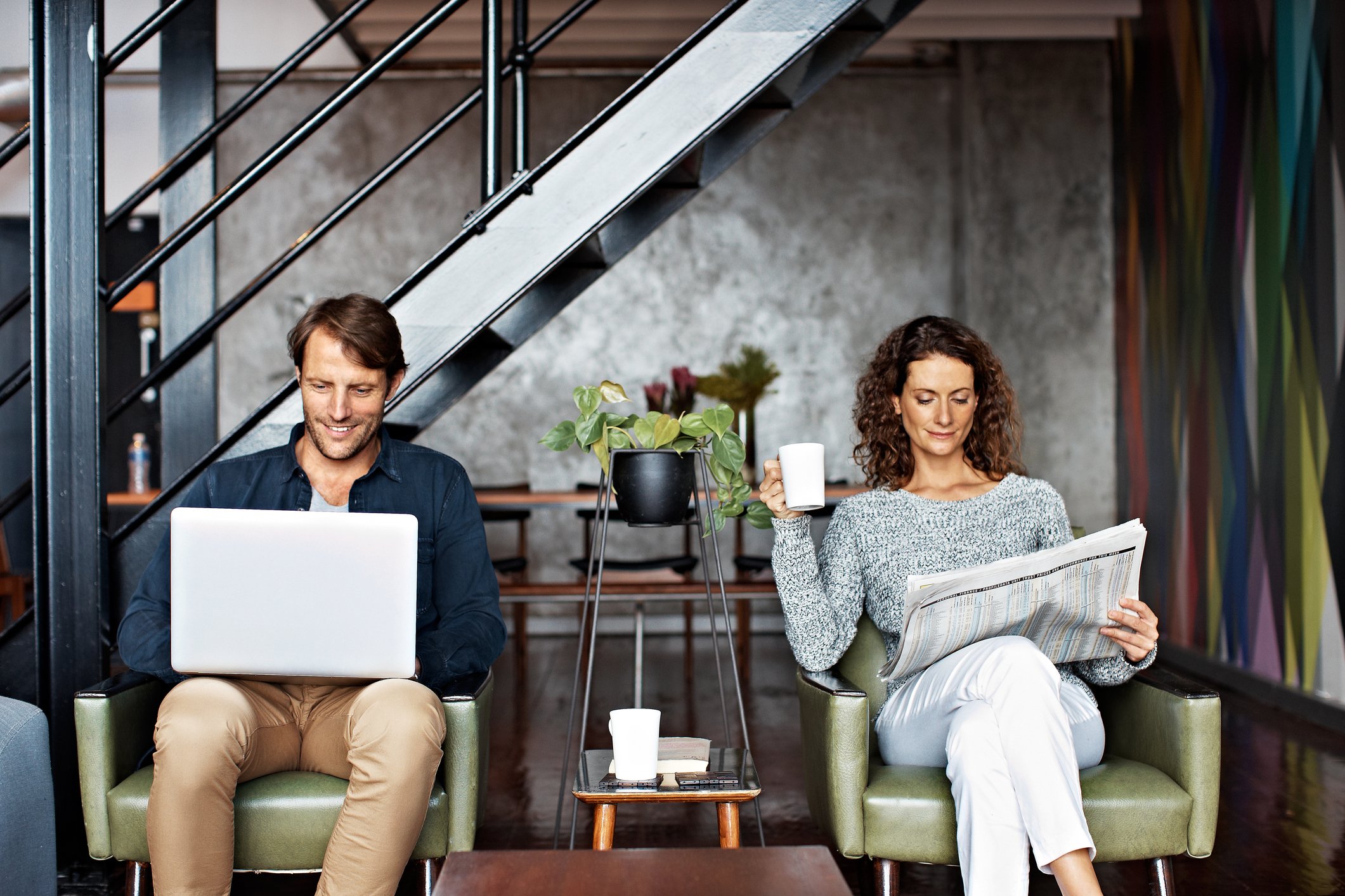 A man reads on his laptop sitting next to a woman reading a print newspaper