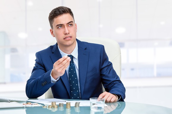 Young businessman in blue suit sitting at desk in office with coin stacks on the desk.