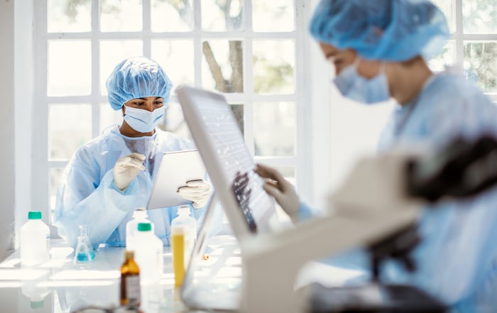 Two lab employees in masks and protective gear use a laptop and a tablet at work.