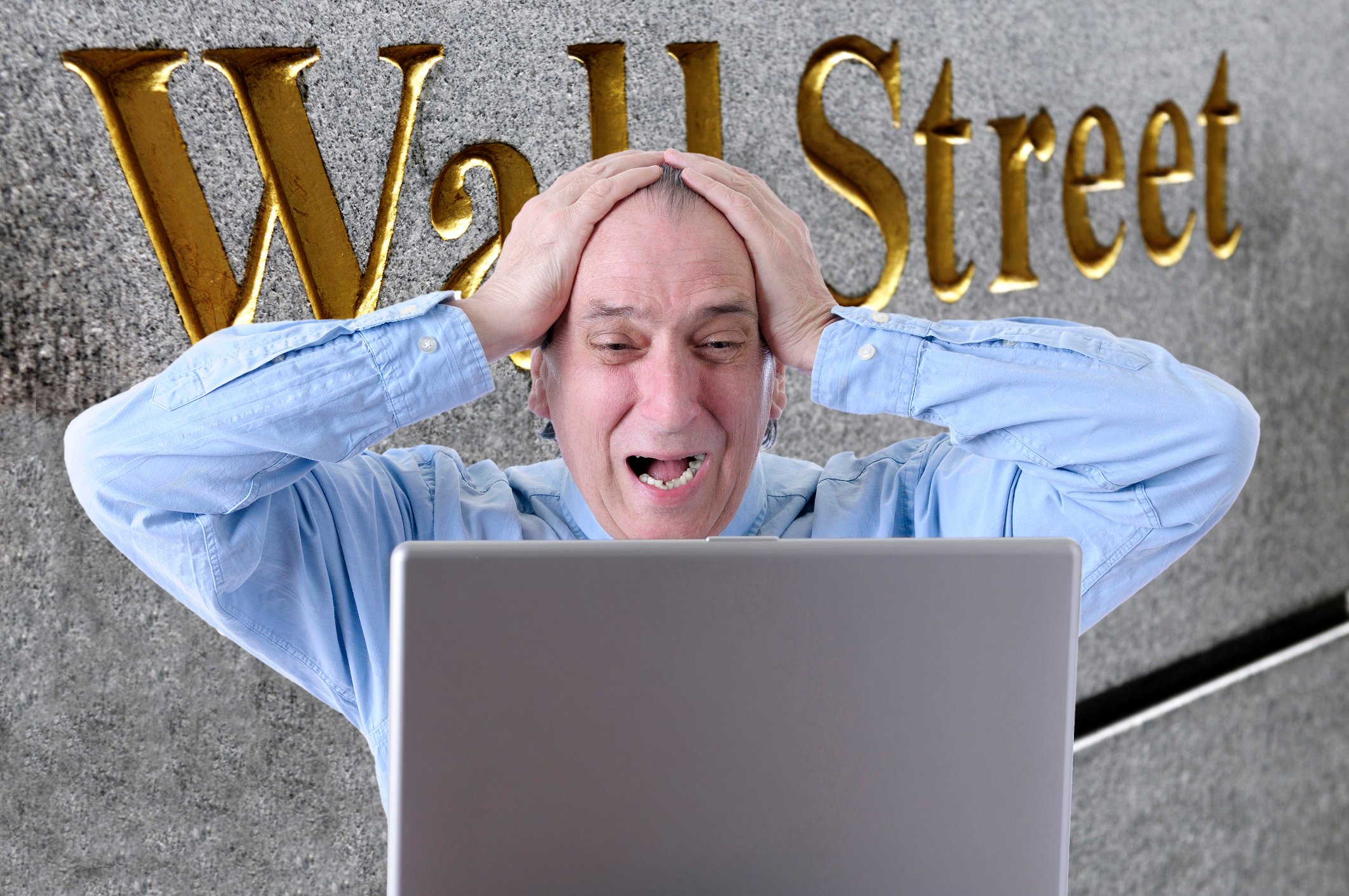 Man with hands on the top of his head and a worried expression looking at a laptop screen with the words Wall Street on a side of a building in the background.