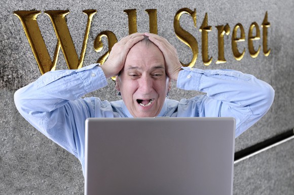 Man with hands on the top of his head and a worried expression looking at a laptop screen with the words Wall Street on a side of a building in the background.