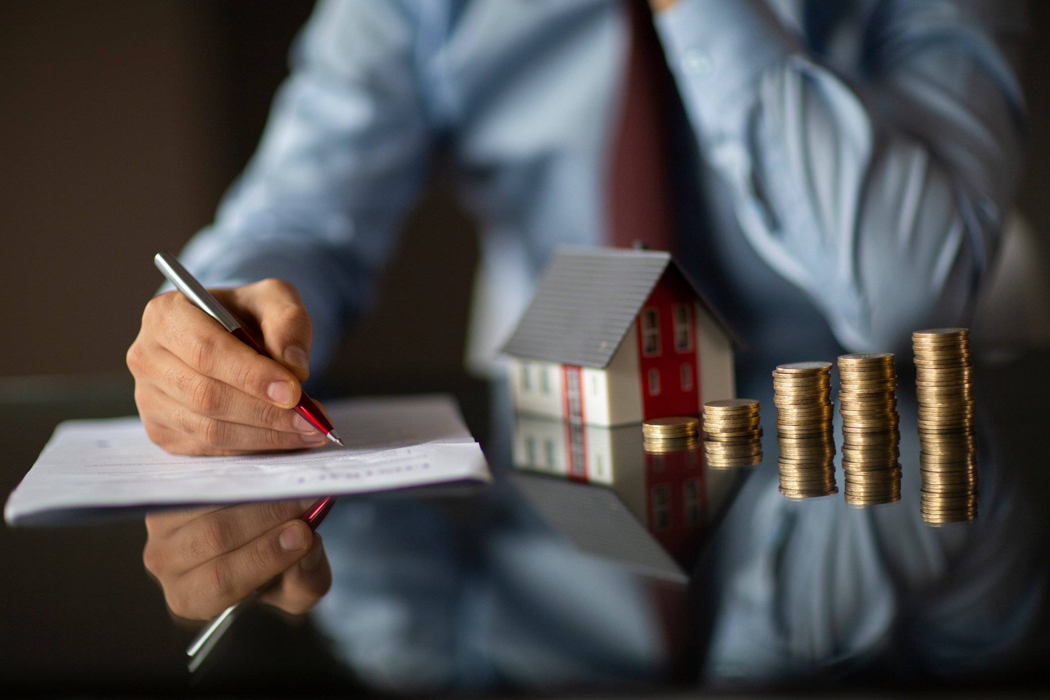 A landlord considers signing a paper on a desktop with a small house and several stacks of coins.