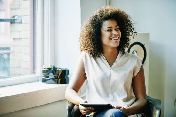 happy young woman sitting in chair near sunny window holding tablet