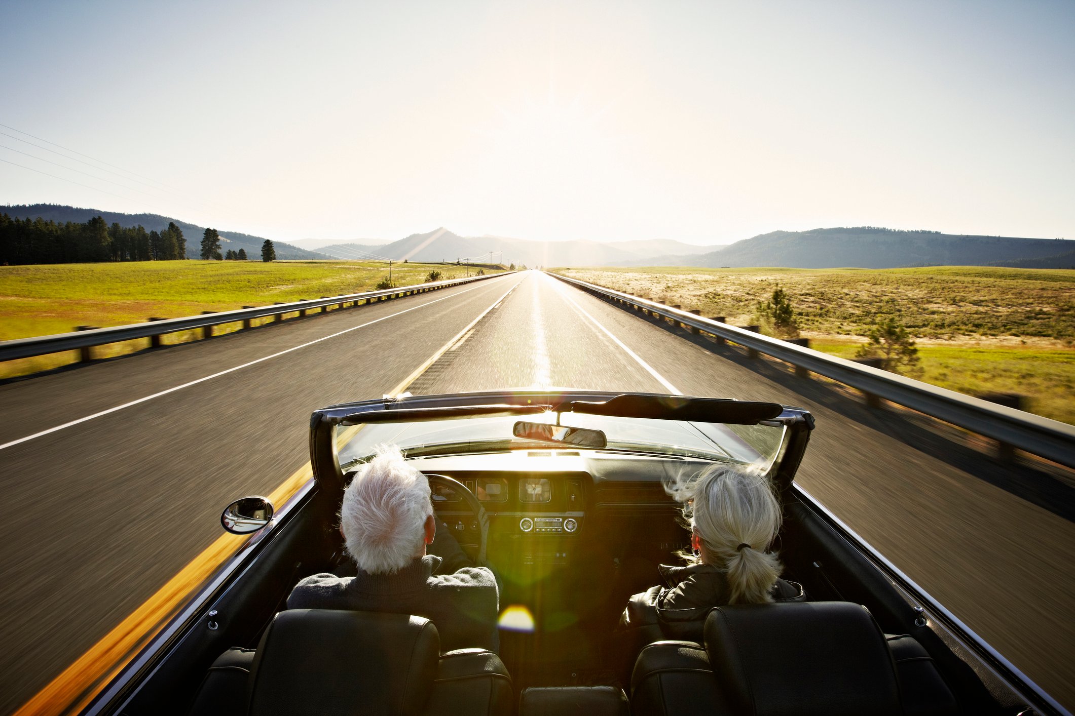 An older couple driving in a convertible down an open road in the countryside.