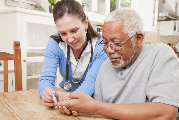 A nurse helping a man at a table take his blood sugar level using a handheld device.