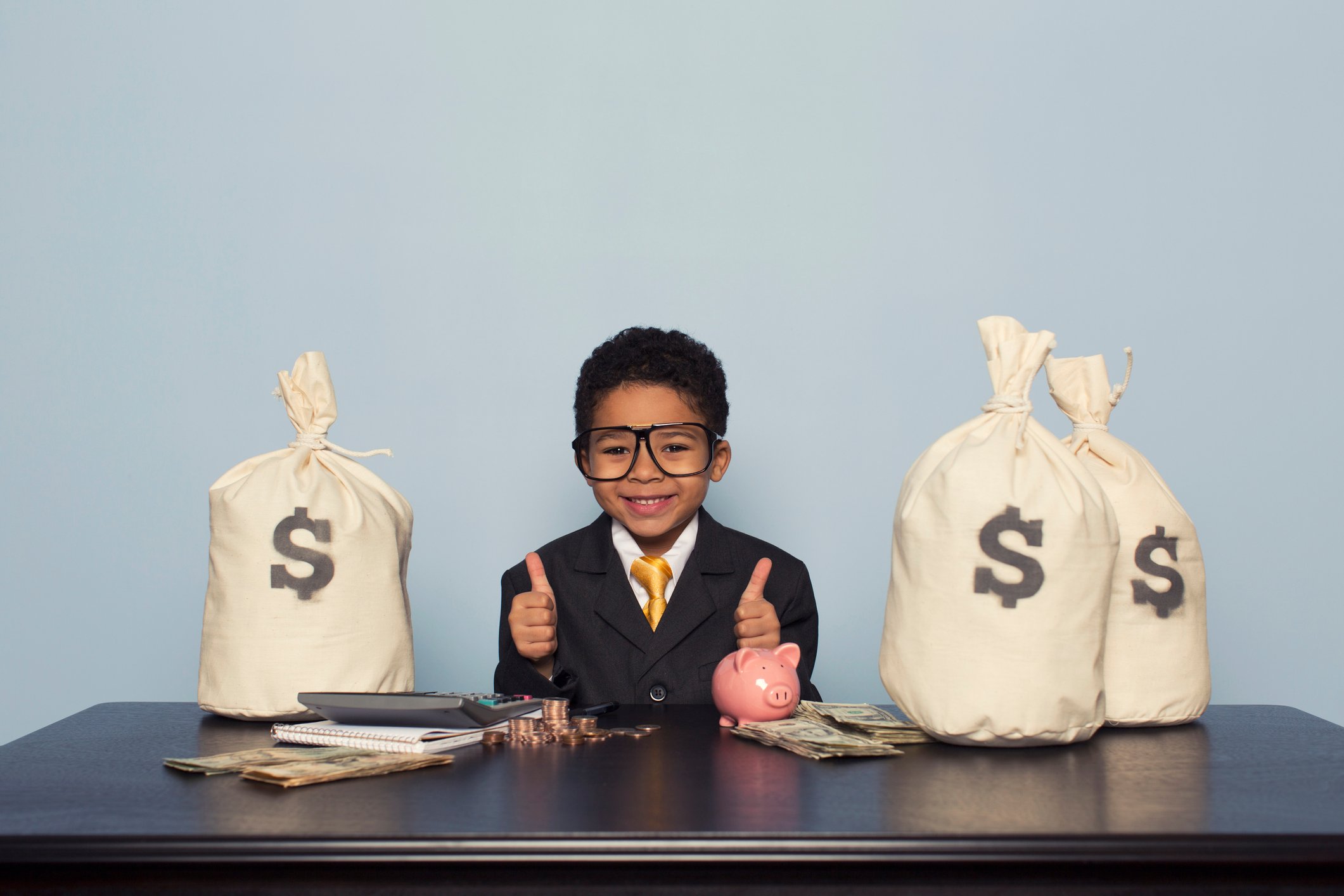 A boy wearing a suit and tie putting his thumbs up as he sits at a table surrounded by money bags.