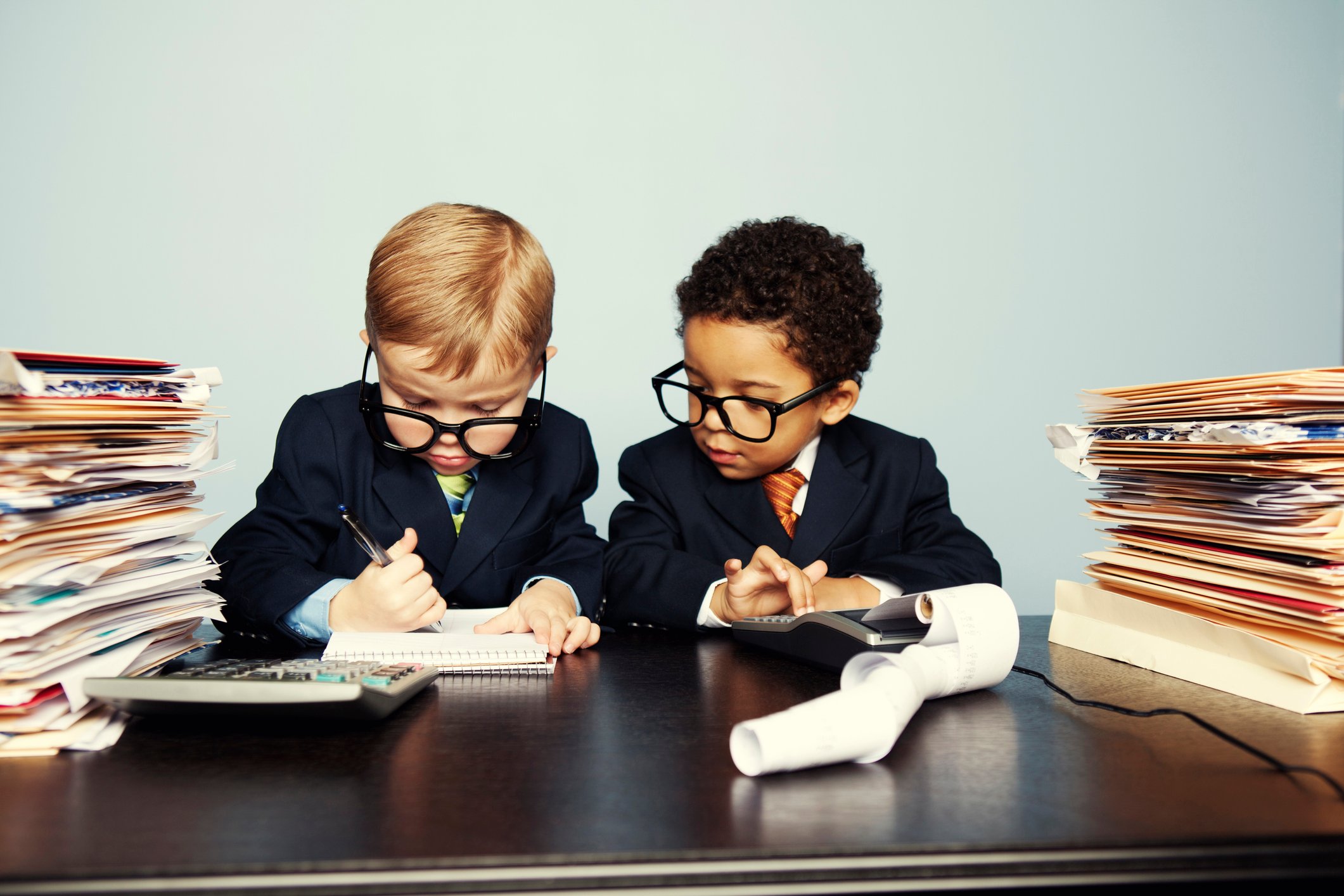 Two toddlers wearing business suits and oversized spectacles appear overwhelmed amid notebooks, a calculator, and stacks of documents in folders.