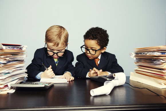 Two toddlers wearing business suits and oversized spectacles appear overwhelmed amid notebooks, a calculator, and stacks of documents in folders.
