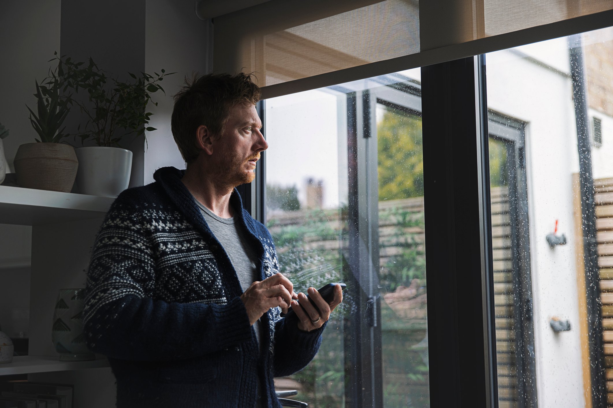 A man, holding a phone, looking out the window, depicting someone looking long-term. 