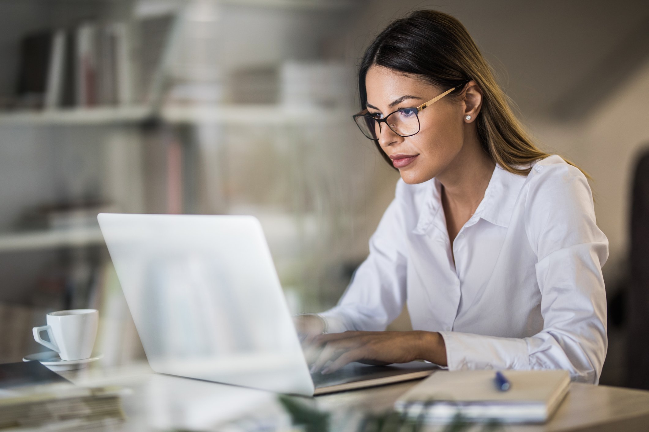 Young adult wearing a collared shirt and glasses types on a laptop with a coffee mug at their side.