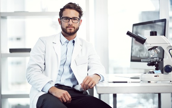 A scientist sits in a laboratory next to a computer and a microscope.
