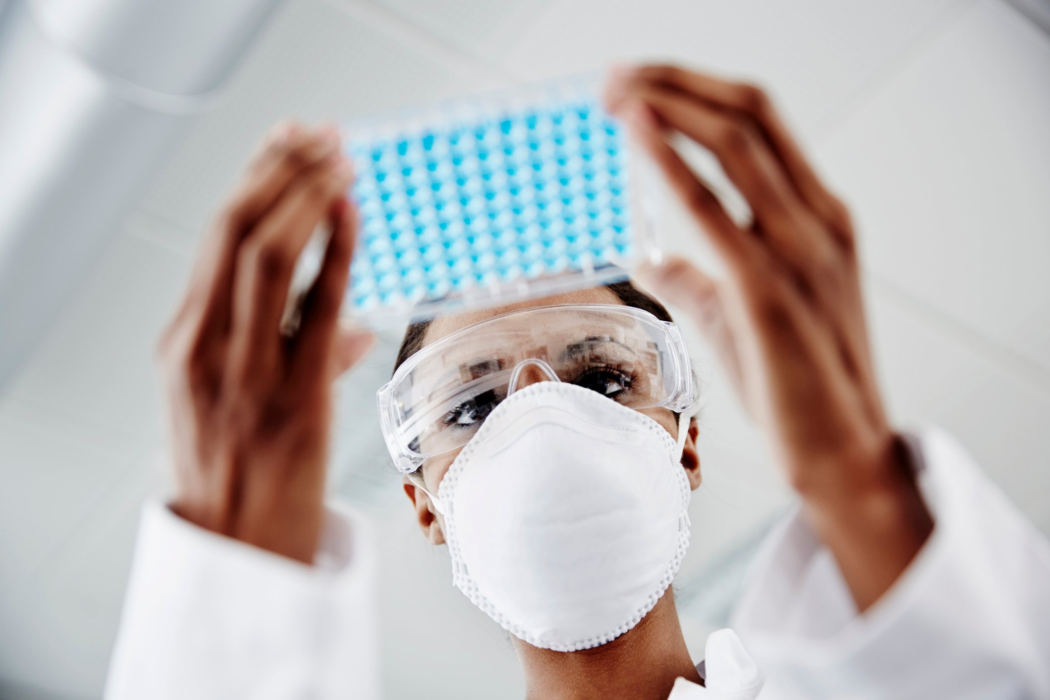 Person in mask, white coat, and goggles holds up a plastic container with blue dots in lab setting.