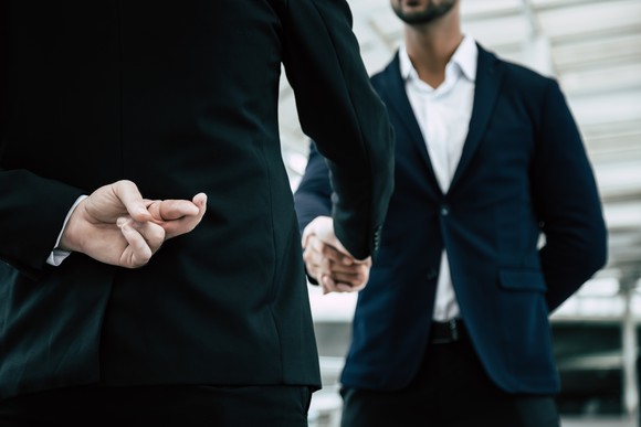 Two businessmen shake hands. One holds two fingers crossed behind his back.