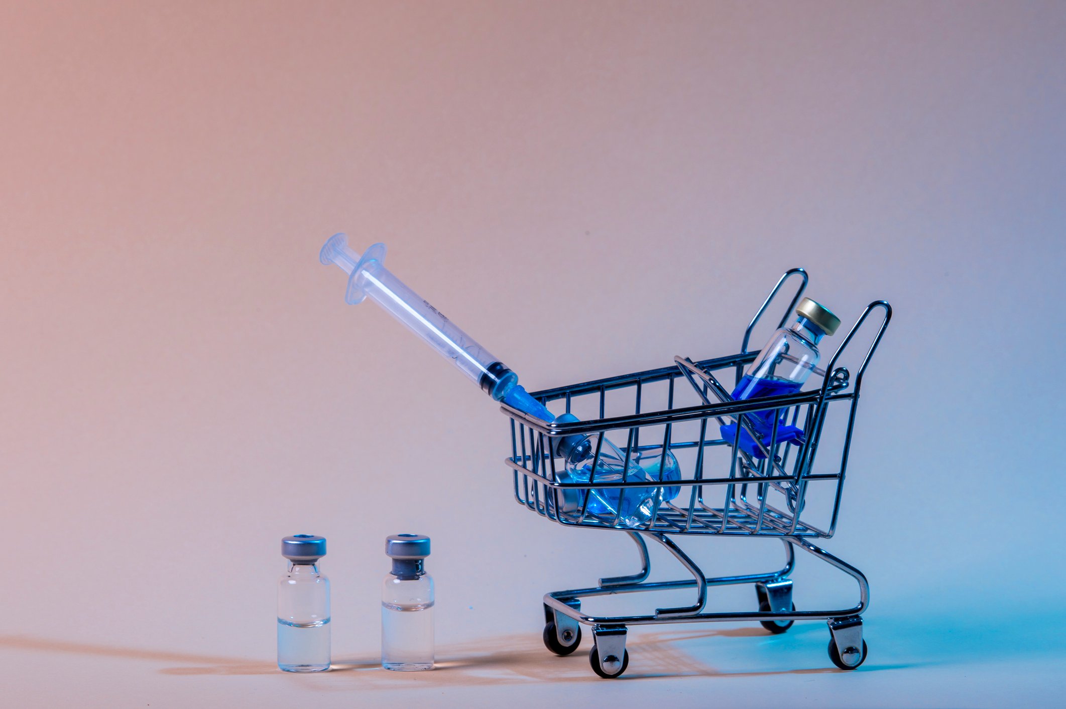 Tiny shopping cart with three vaccine vials and a syringe in it and two vaccine vials next to the cart