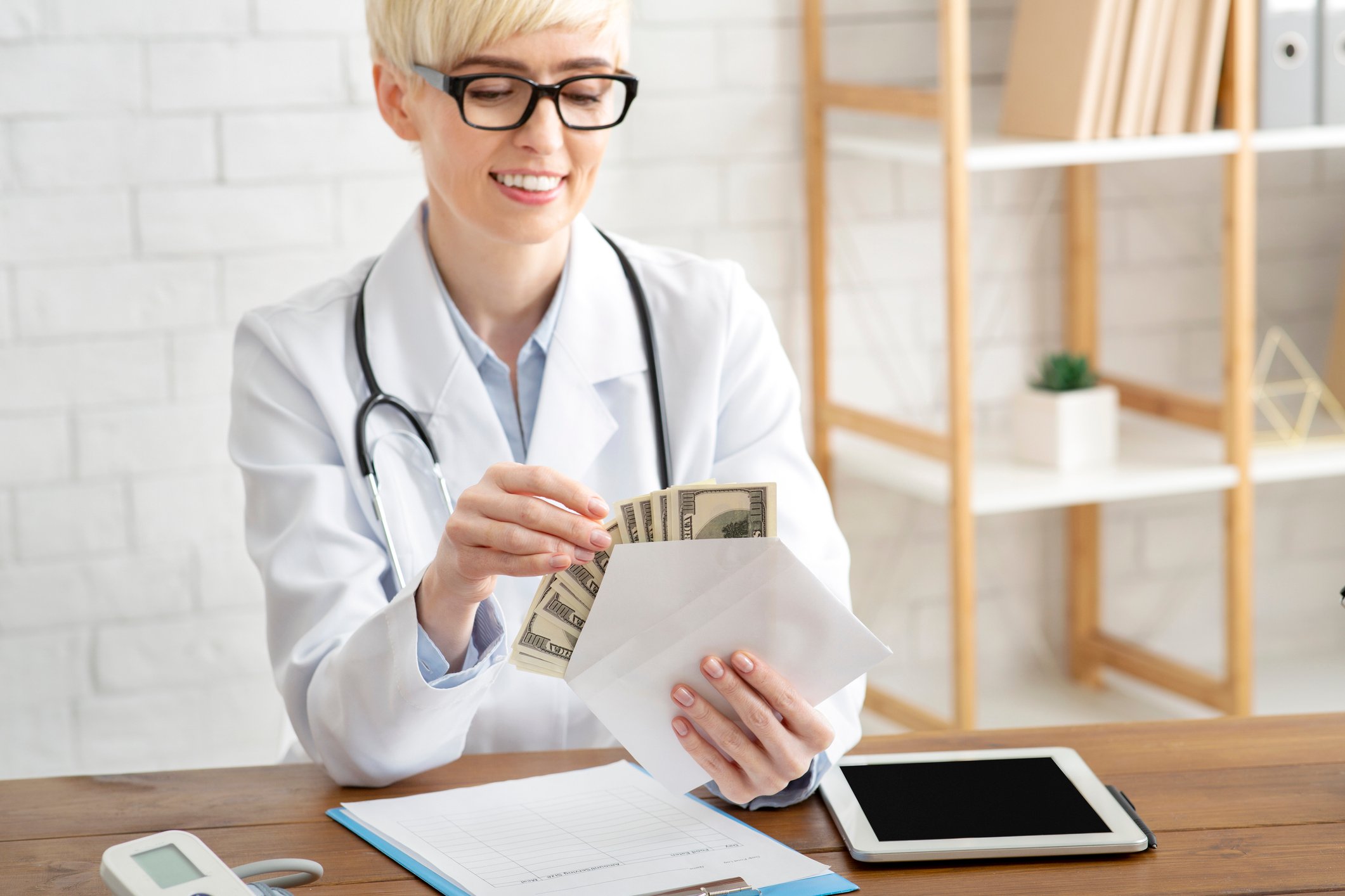 Smiling doctor sitting at a table, holding an envelope filled with cash.