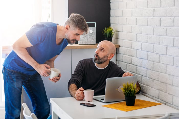 A man sitting at his laptop discusses something with a man standing next to him.