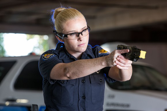 A female police officer holds a TASER weapon as if preparing to shoot it with a police vehicle in the background