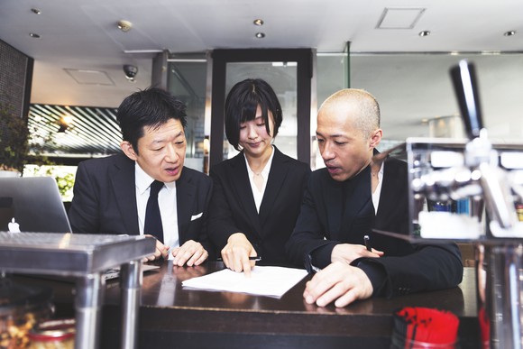 Three people ponder signing a contract on a table in front of them.