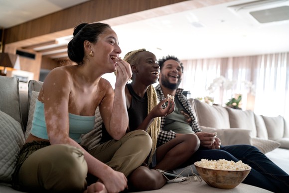 Three people sitting on a couch while sharing a bowl of popcorn and laughing.
