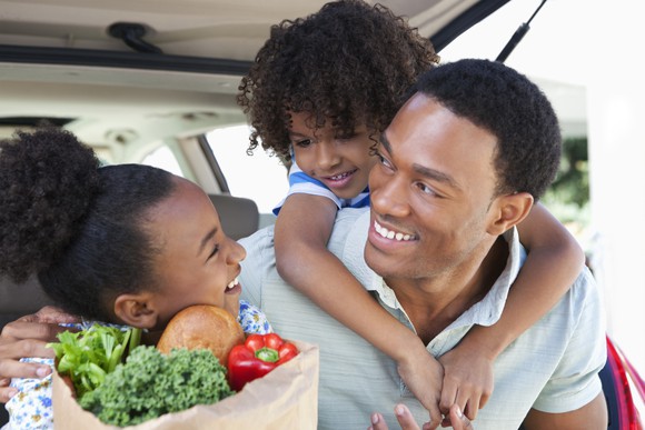 A man and two children hold a grocery bag near a car trunk.