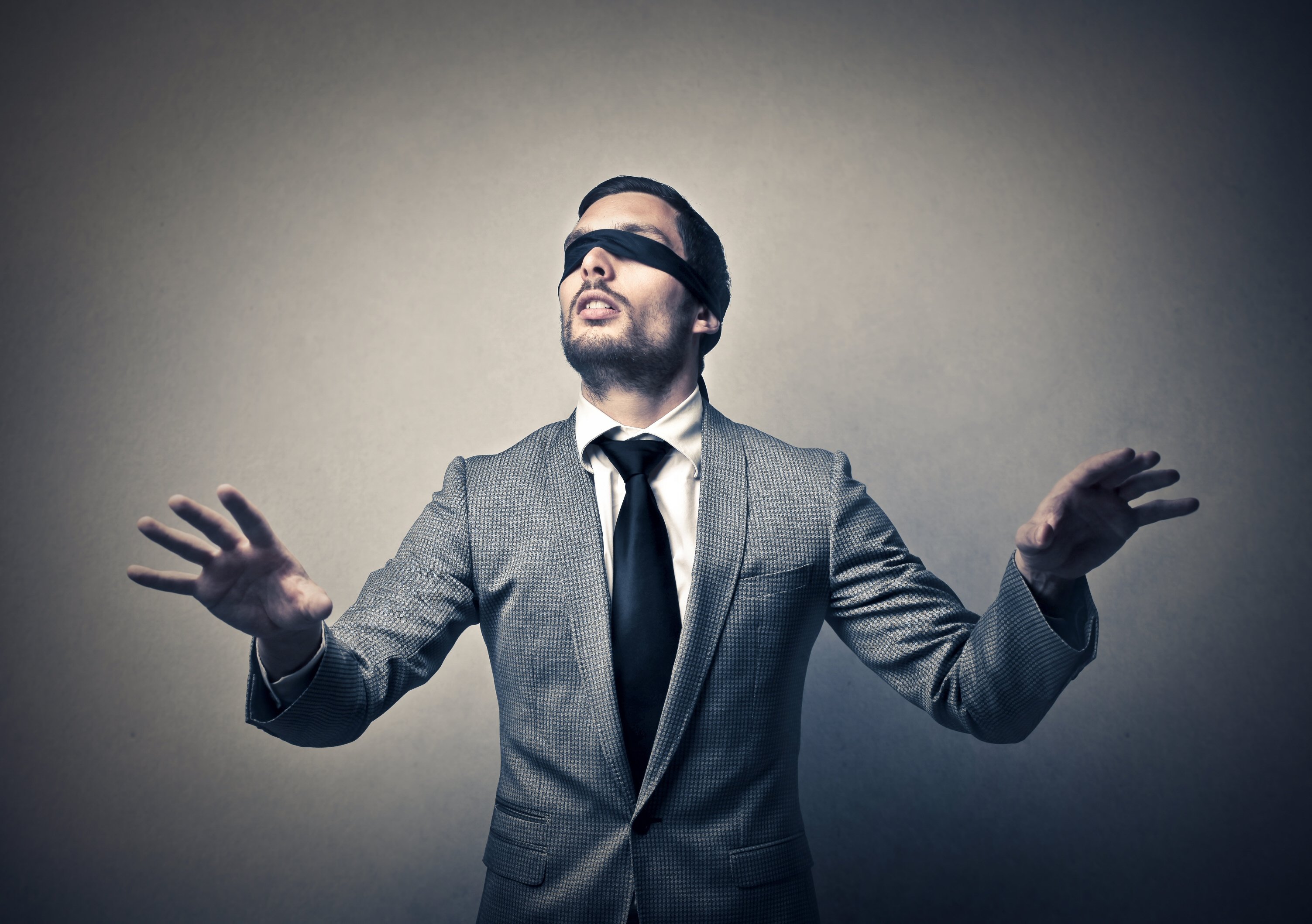 A man in a suit wears a blindfold and holds his hands up.